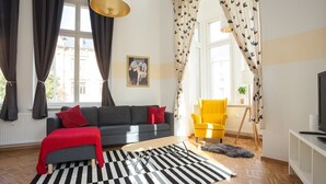 Ein Wohnzimmer mit grauem Sofa, roter Decke, Teppich in Schwarzweiß-Streifen, gelbem Sessel und einem Fernseher auf einem weißen Schrank.