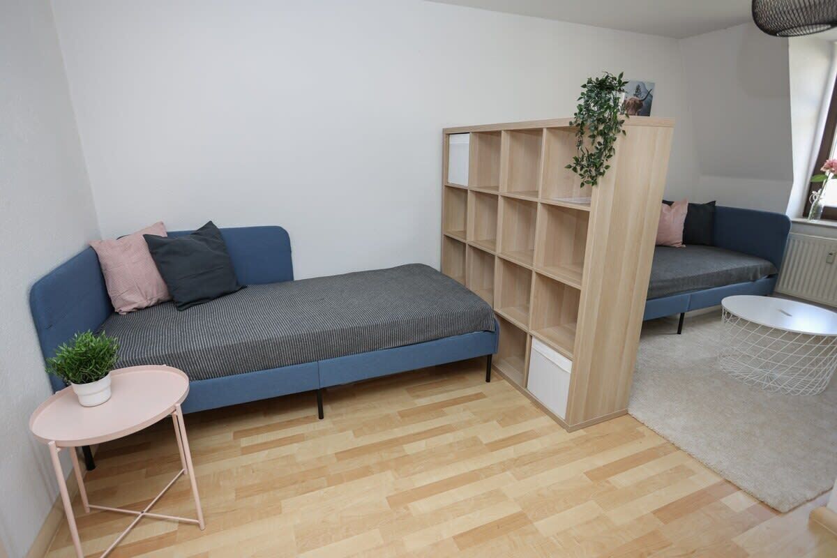 En kompakt opholdsstue med en blå sofa, et lille sidebord med en plante, en træbogreol og et rundt hvidt bord.