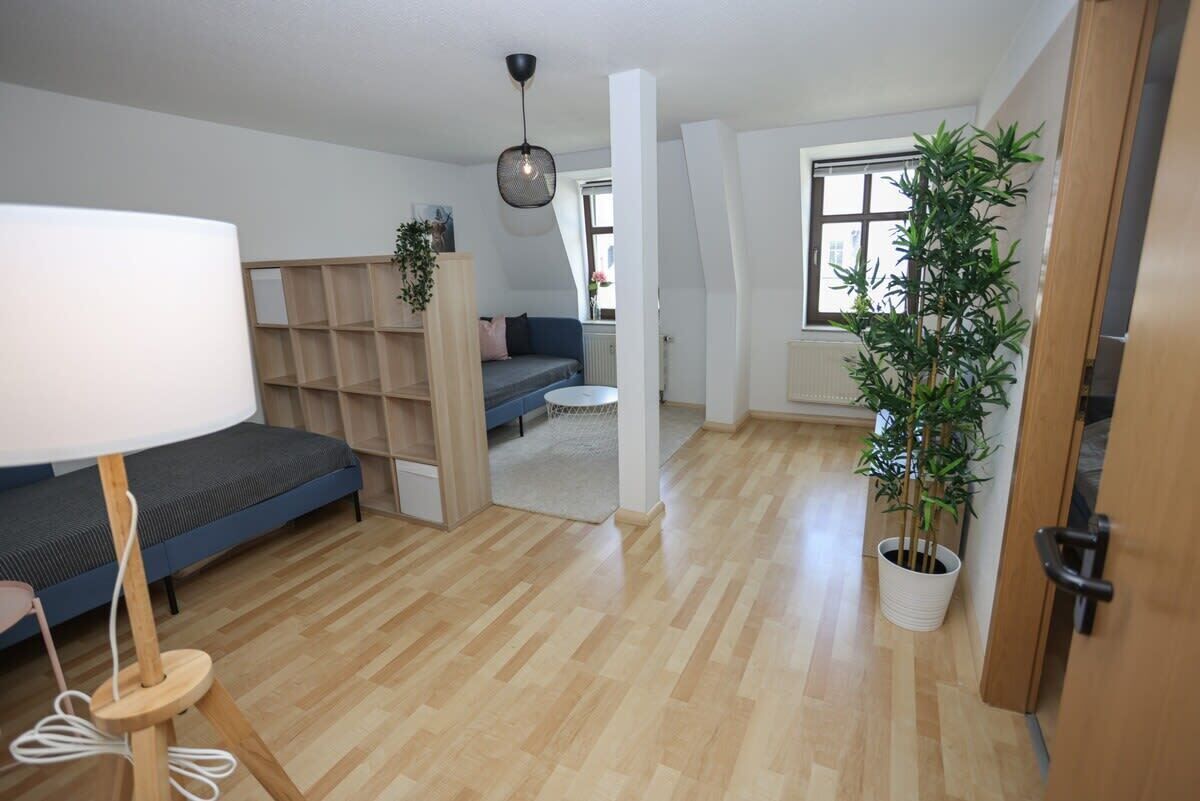 Ein moderner, offener Wohnbereich mit Holzboden, einer Couch, einer Lampe, einem Bücherregal und einem Blumentopf.