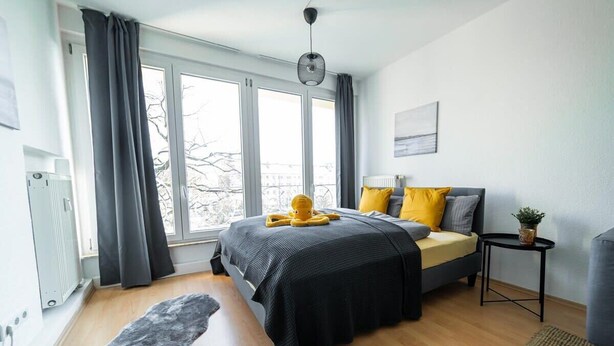 A modern bedroom with a large bed, a bedside table, a radiator, and a window with curtains. - FullHouse | Chemnitz | HS46 | 3 Bedroom (Chemnitz)