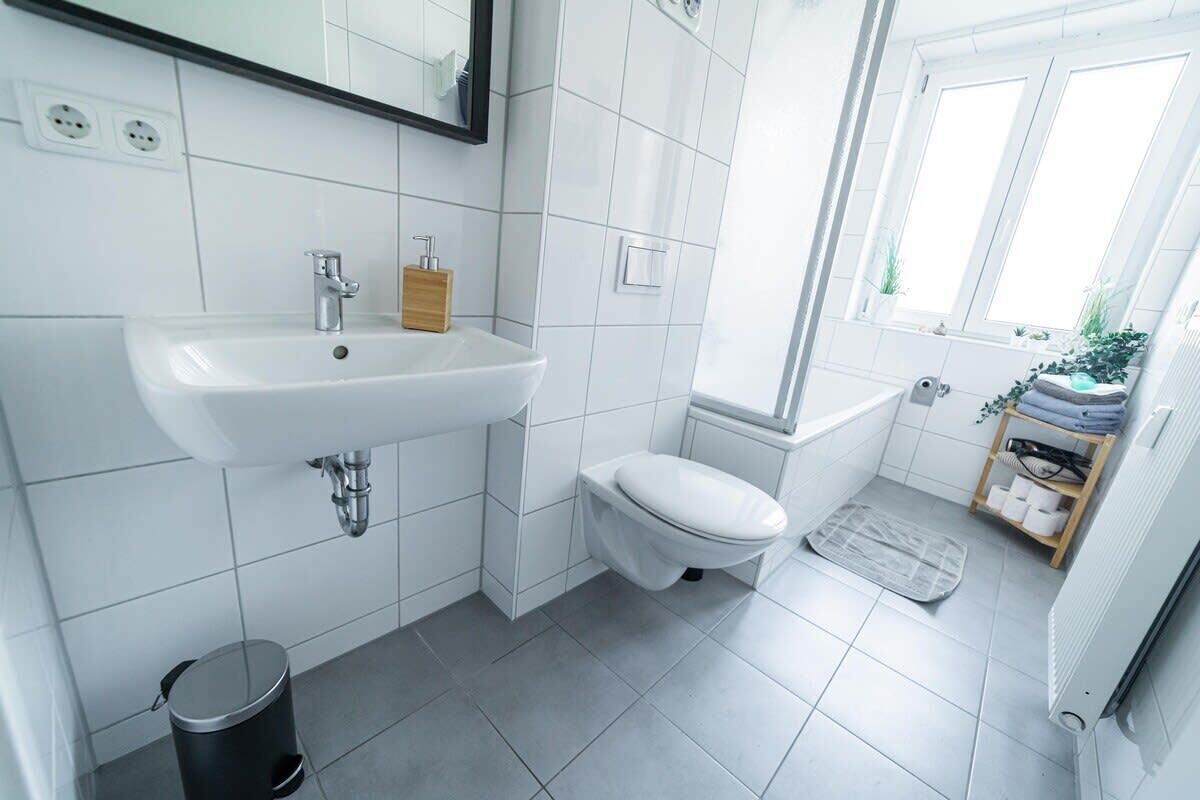 Un baño moderno con un lavabo, inodoro y ducha blancos. Hay un pequeño cubo de basura y una ventana con plantas.