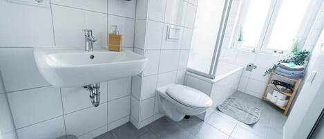 Un baño moderno con un lavabo, inodoro y ducha blancos. Hay un pequeño cubo de basura y una ventana con plantas.