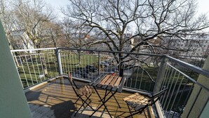 A balcony with a wooden deck, metal railing, and a view of a park and buildings in the background. - FullHouse | Chemnitz | HS46 | 3 Bedroom (Chemnitz)