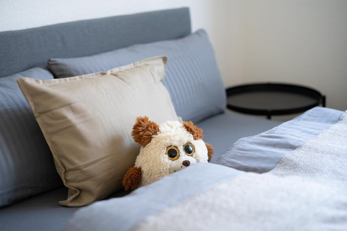 A bed with a grey headboard, a beige pillow, and a plush toy peeking out from under the covers.