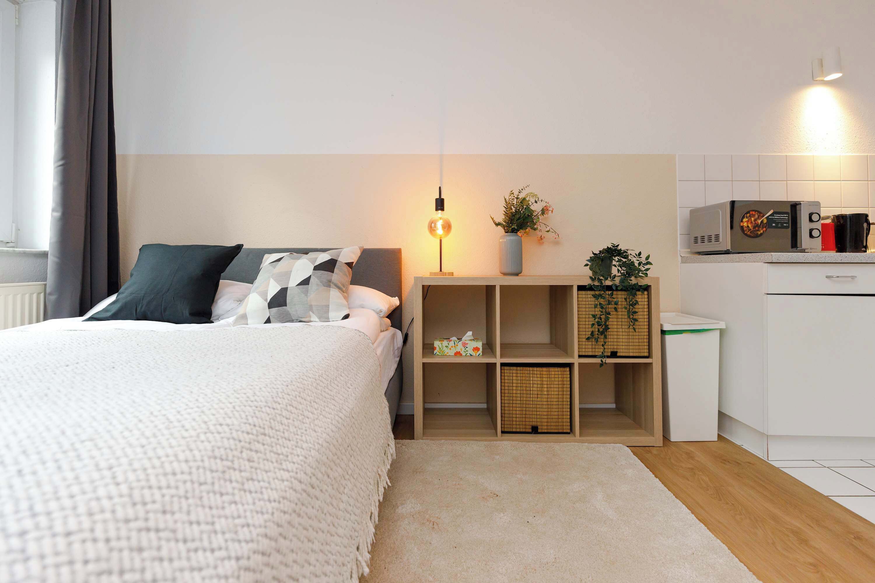 A modern bedroom with a bed, bedside table, lamp, and a small kitchenette area with a microwave and coffee maker.