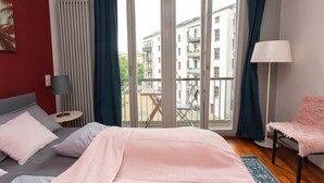 Ein Schlafzimmer mit einem Bett, einem Sessel, einer Lampe, einem Fenster und Blick auf Gebäude.