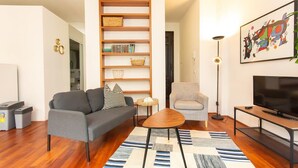 Ein modernes Wohnzimmer mit einem grauen Sofa, einem Holzregal, einem kleinen runden Tisch, einem grauen Sessel, einem Fernseher auf einem Schrank und einem farbenfrohen abstrakten Gemälde an der Wand.