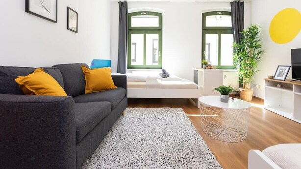 A modern living room with a grey sofa, yellow cushions, a white coffee table, and a large window with green curtains. - FullHouse | Halle | R6 Amsterdam (Halle (Saale))