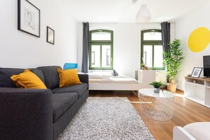 A modern living room with a grey sofa, yellow cushions, a white coffee table, and a large window with green curtains. - FullHouse | Halle | R6 Amsterdam (Halle (Saale))
