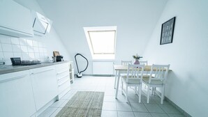 A modern kitchen with white cabinetry, a dining area with white chairs, a window, and a radiator. - FullHouse | Magdeburg | L10-4 | Loft-Flair (Magdeburg)
