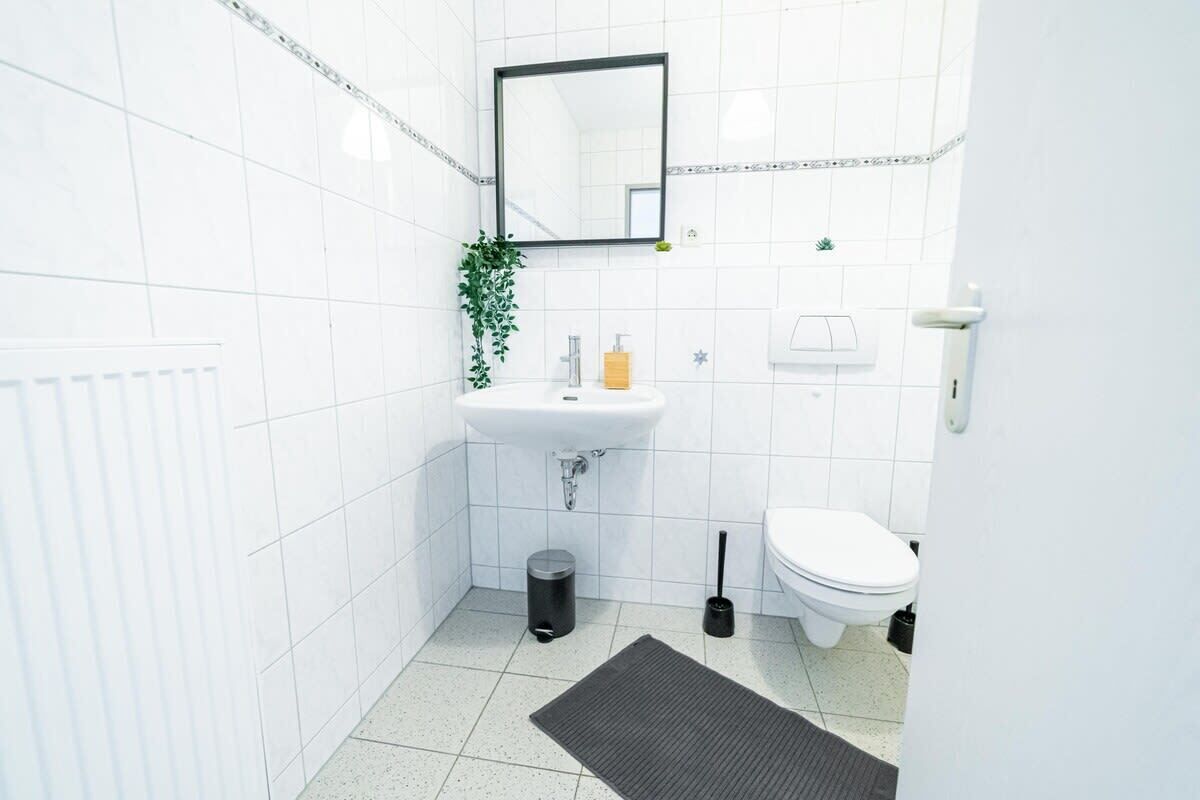 Un bagno piccolo e pulito, con piastrelle bianche, un lavandino bianco, un water, uno specchio, una pianta e un cestino.