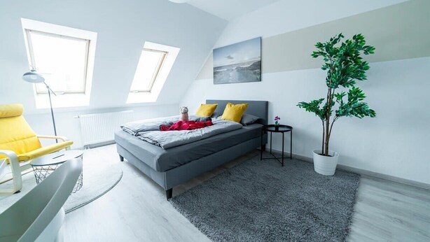 A modern bedroom with a bed, a chair, a small table, a potted plant, and two windows. - FullHouse | Magdeburg | L10-4 | Loft-Flair (Magdeburg)