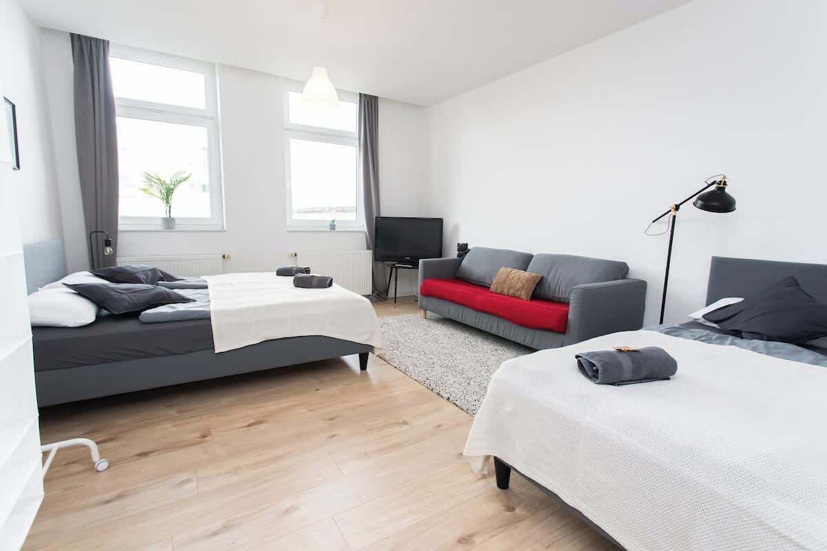 Una habitación de hotel moderna con dos camas, un sofá gris, un televisor y una lámpara.