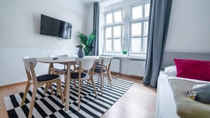 A modern room with a dining table and chairs, a flat-screen TV mounted on the wall, a large window with curtains, and a bed with a red pillow. - FullHouse | M18-4 | Kings Palace (Erfurt)