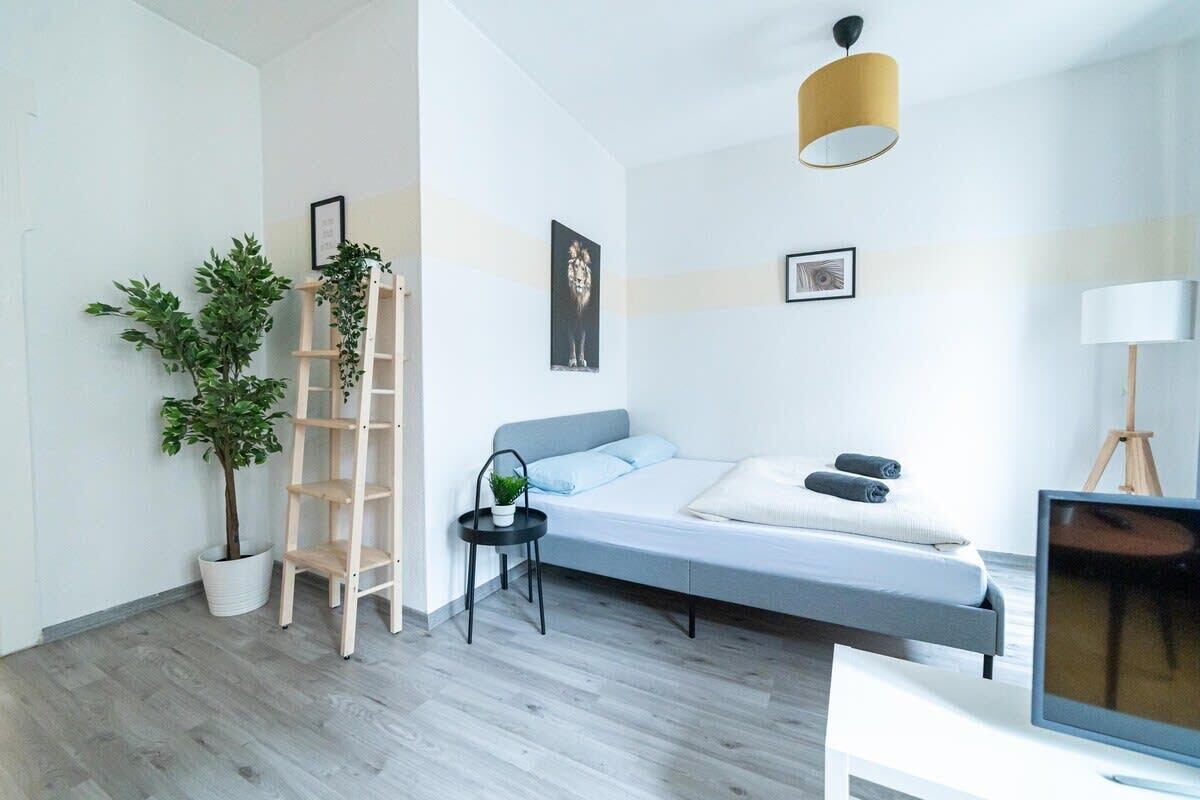 Ein modernes Schlafzimmer mit einem Bett, einem kleinen Schreibtisch, einem Fernseher, einer Pflanze und einer an der Wand stehenden Leiter.