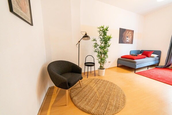 Un salon moderne avec un tapis rond en fibres naturelles, un fauteuil foncé, un canapé gris agrémenté de coussins rouges, un lampadaire noir, un pot de bambou et un tableau encadré représentant un lion, accroché au mur.