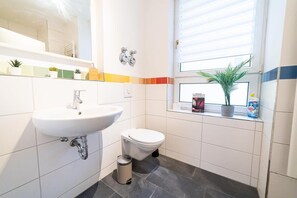 Une salle de bain moderne avec un lavabo blanc, des toilettes et une petite poubelle. Il y a une fenêtre avec des stores et une étagère contenant des articles de toilette.