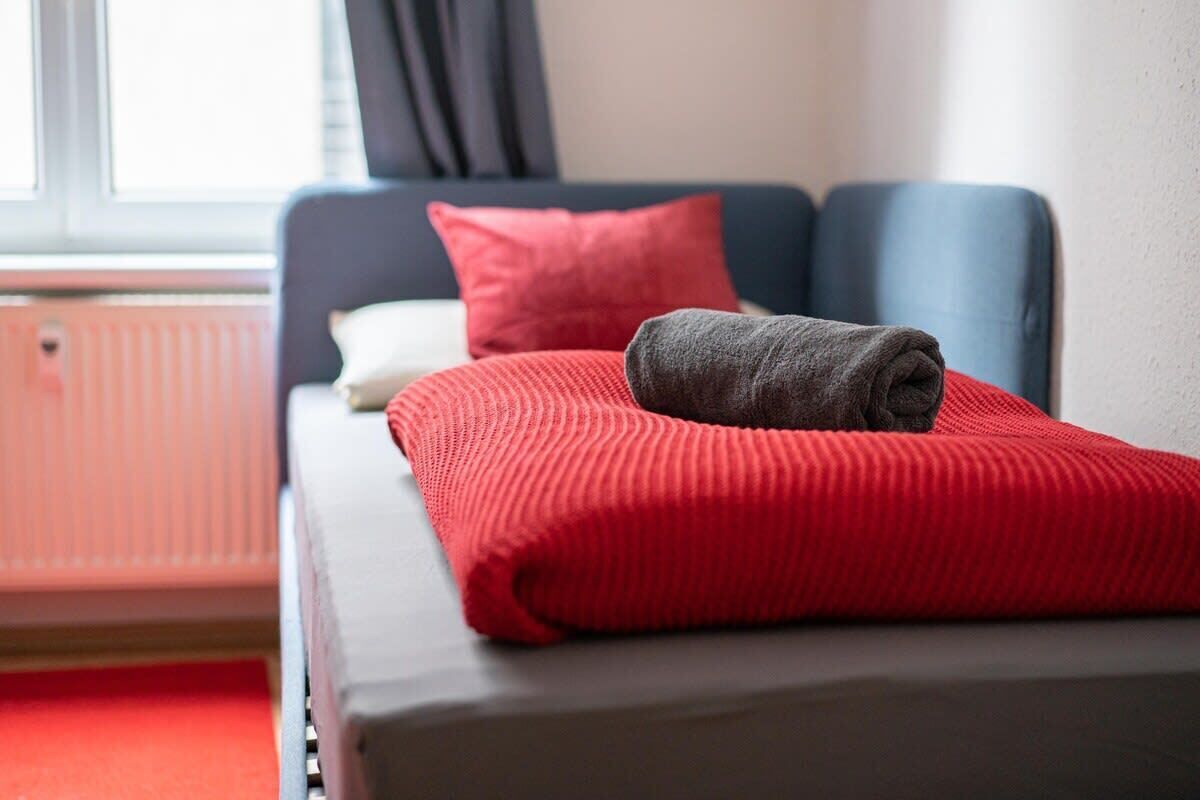 A neatly made bed with a red blanket and a folded gray towel.