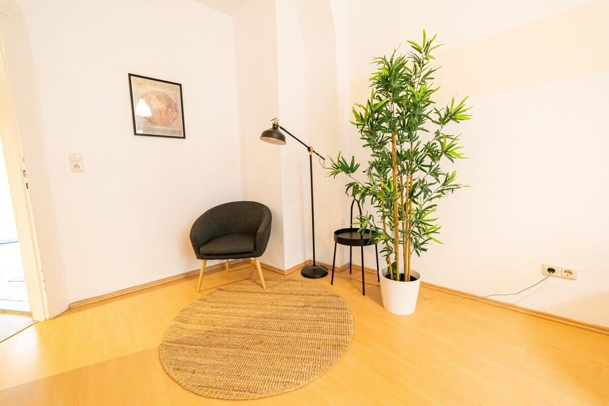 A room with a wooden floor, a black chair, a round woven rug, a tall bamboo plant in a white pot, a black floor lamp, and a framed picture on the wall.