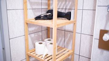 Une étagère en bois avec du papier toilette et un sèche-cheveux noir, dans une salle de bain aux murs carrelés.