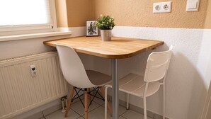 Ein kleiner Essbereich mit einem Holztisch und zwei weißen Stühlen. Auf dem Tisch steht ein Topfpflanze, und darunter ein Heizkörper.