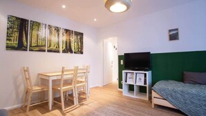 A room with a bed, a TV, a desk with chairs, and three forest-themed wall art pieces.