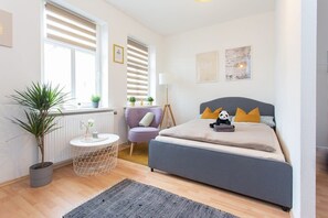 A bedroom with a bed, a chair, a small table, a plant, and a window with blinds. - FullHouse | Chemnitz | S35 Little Sun| Studio (Chemnitz)