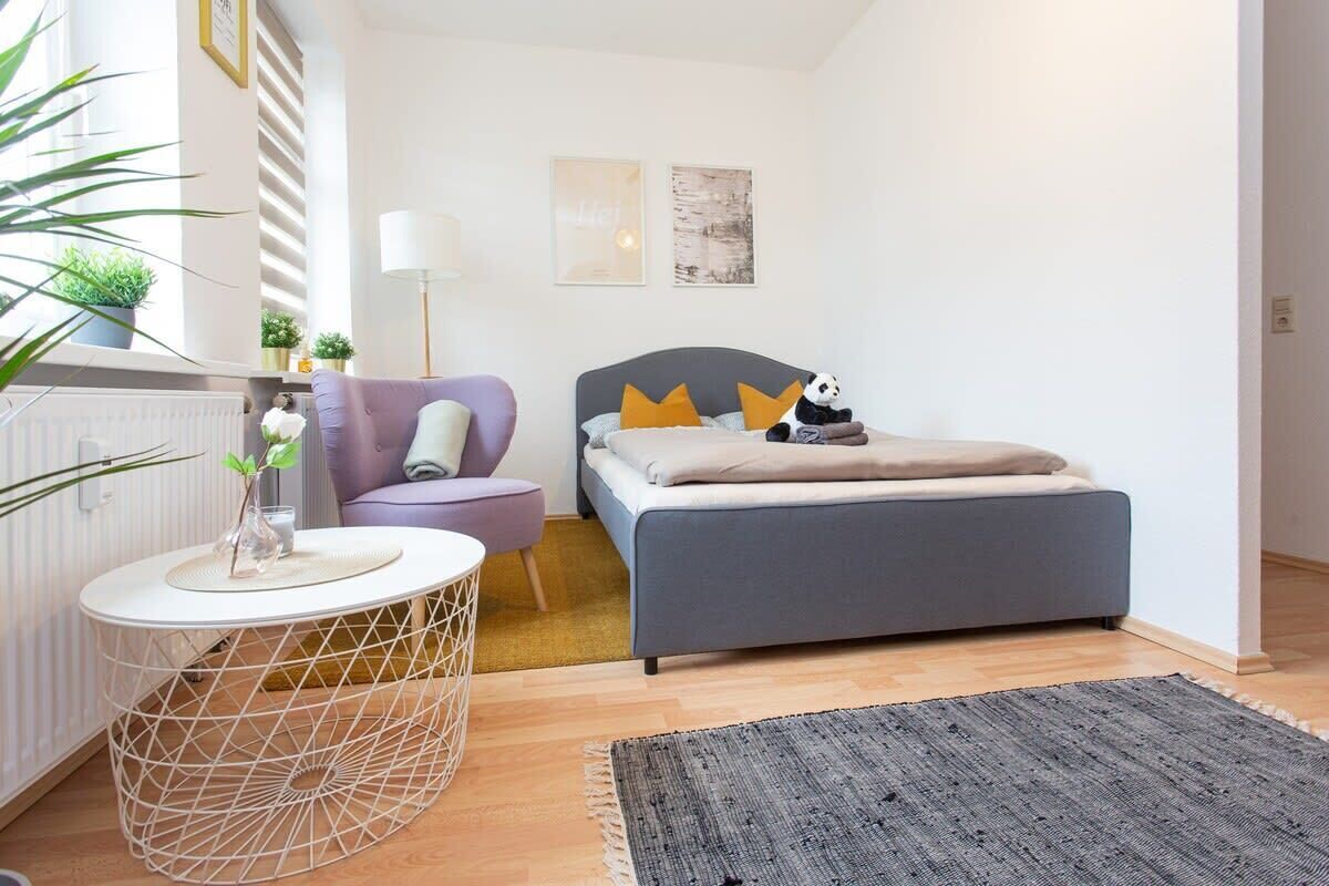 Un dormitorio moderno con una cama gris, una mesita redonda de base blanca, un sillón púrpura y una alfombra gris.