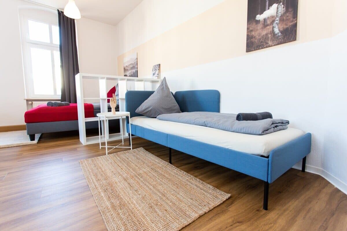 A modern bedroom with a blue sofa bed, a red sofa, a white side table, and a woven rug.