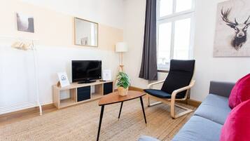 Un salon moderne avec un canapé, un fauteuil, un téléviseur, une petite table sur laquelle est posée une plante, et une fenêtre avec des rideaux.