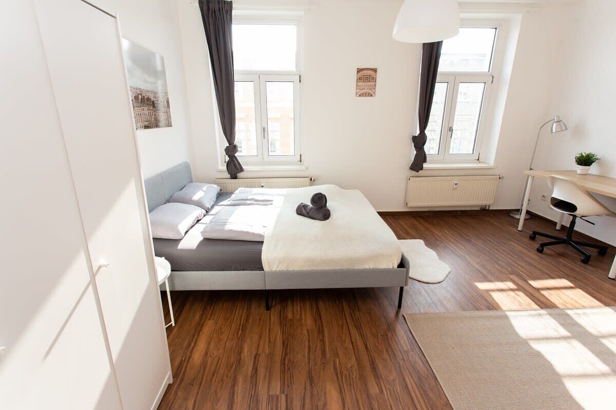 A modern bedroom with a bed, a desk, and a chair. There are two windows with curtains, a radiator, and a small plant on the desk.