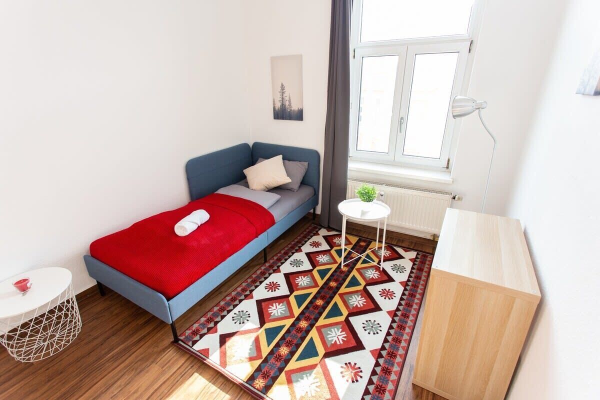 A small, well-lit room with a bed, a small table, a radiator, and a window.