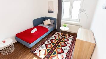 A small, well-lit room with a bed, a small table, a radiator, and a window.