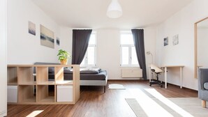 Una camera da letto moderna e luminosa, con un letto, una scrivania e una sedia. Sono presenti due finestre con tende, un termosifone e una pianta in vaso.