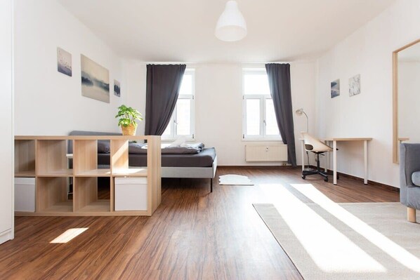 A modern, well-lit bedroom with a bed, a desk, and a chair. There are two windows with curtains, a radiator, and a potted plant.