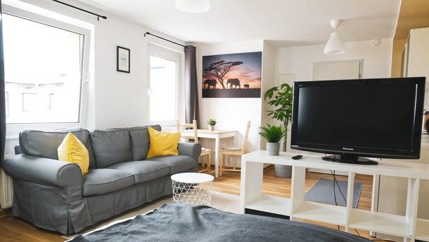 A modern living room with a grey sofa, a flat-screen TV, and a dining area with a small table and chairs. - FullHouse | Halle | T28 Elephant Apt. (Halle (Saale))