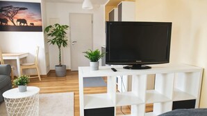 A modern living room with a flat-screen TV on a white console, a dining area with a table and chairs, a potted plant, and a painting of elephants on the wall. - FullHouse | Halle | T28 Elephant Apt. (Halle (Saale))