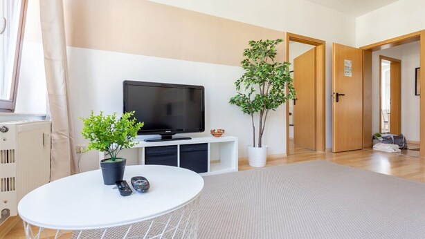 Een moderne woonkamer met een flatscreen televisie, een ronde witte tafel, twee potplanten en een groot vloerkleed.