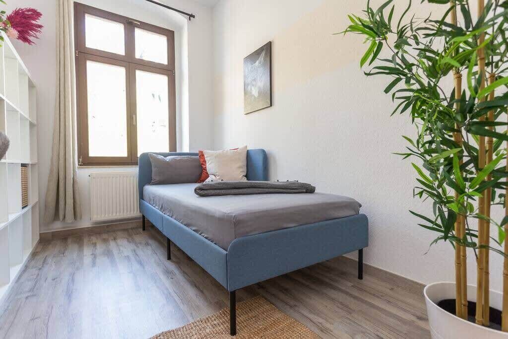Un dormitorio pequeño con una cama individual, una ventana con cortinas, un radiador, un cuadro en la pared y una planta en maceta.