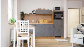 A compact kitchen with grey cabinets, a microwave, and a built-in oven. There's a white chair and a small dining table with a plant beside it. - FullHouse | K70 Goethe | Balcony | Washing Machine (Eisenach)