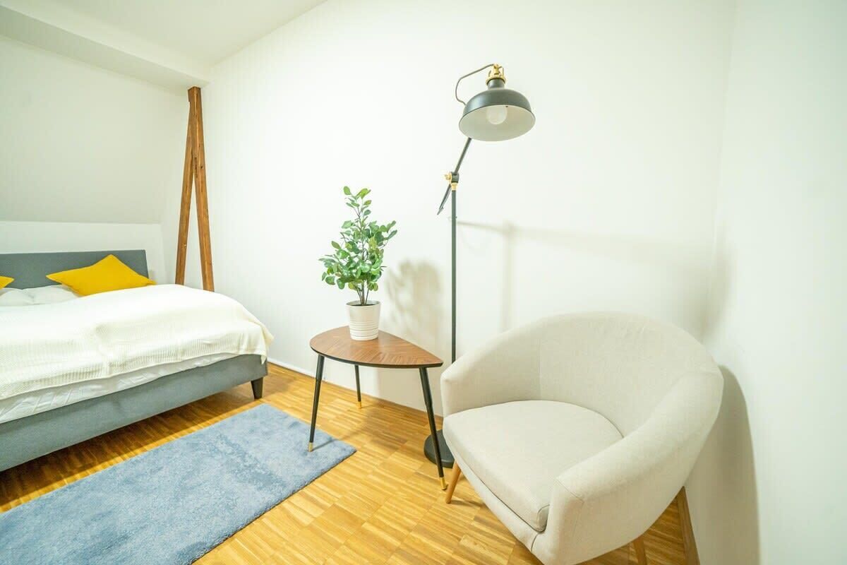 A bedroom with a bed, a small round table with a plant, a floor lamp, and a white armchair.