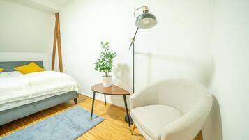 A bedroom with a bed, a small round table with a plant, a floor lamp, and a white armchair.