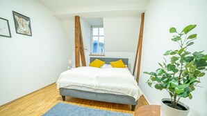 A bedroom with a bed, a blue rug, a potted plant, and a window.