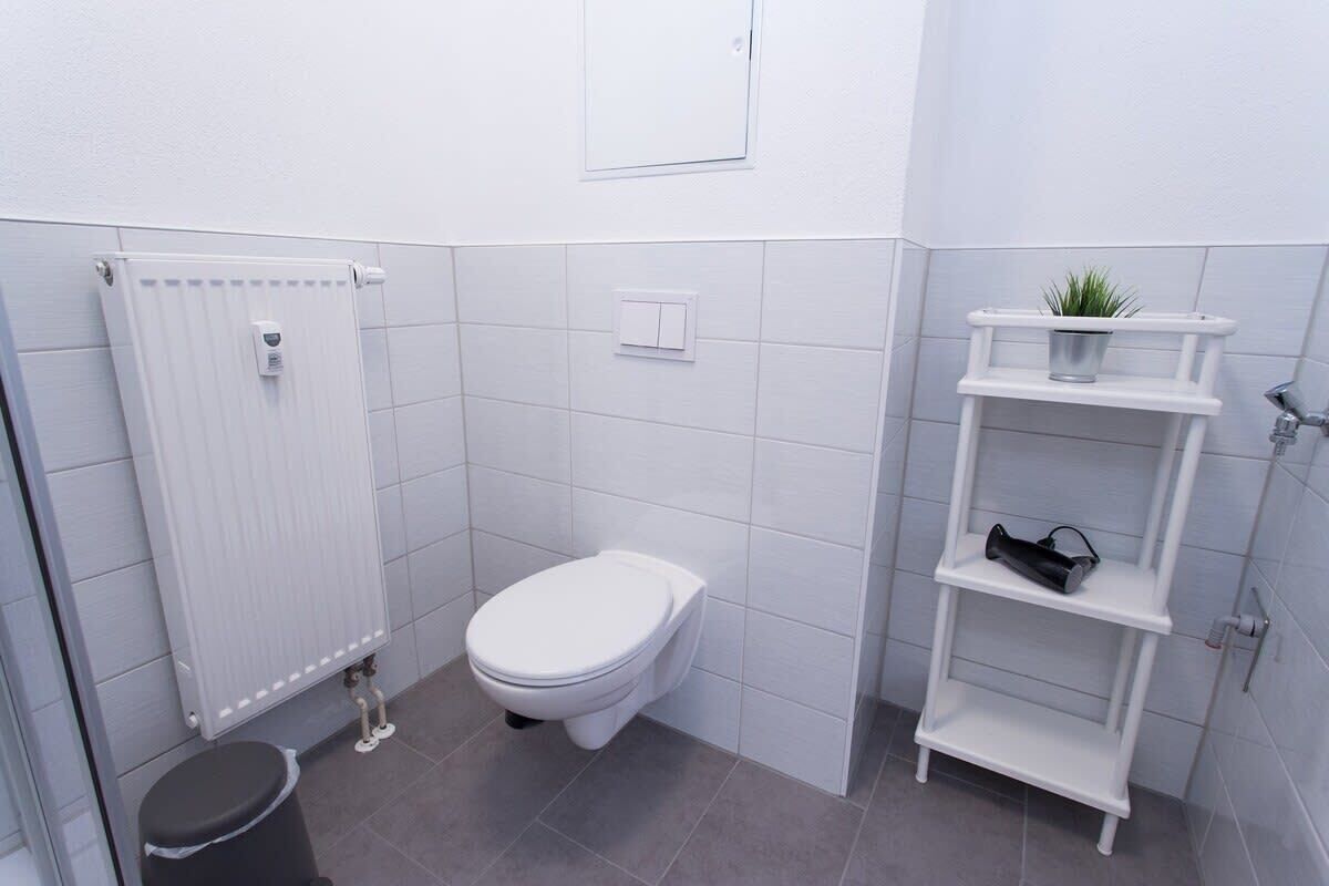 Une petite salle de bain blanche, équipée de toilettes, d’un radiateur, d’une poubelle et d’une étagère sur laquelle est posée une plante.