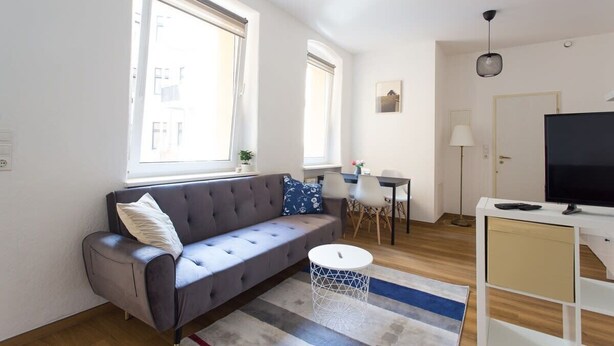 A modern living room with a grey sofa, a round coffee table, and a TV on a white stand. There are two windows providing natural light and a small table with chairs in the background. - FullHouse | Halle | T28 Hawk Apt. (Halle (Saale))