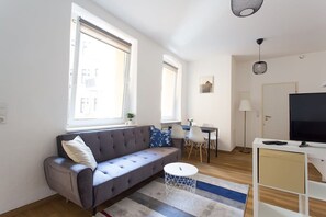 Un salon moderne avec un canapé gris, une table basse ronde et un téléviseur posé sur un meuble blanc. Deux fenêtres laissent entrer la lumière naturelle, et on aperçoit au fond une petite table entourée de chaises.