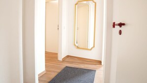 Un couloir doté d’un plancher en bois, d’un tapis sombre, d’une porte blanche avec une poignée rouge et d’un miroir encadré d’or accroché au mur.