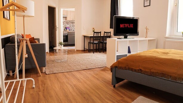 Une chambre d’hôtel moderne équipée d’un lit, d’un téléviseur diffusant « NETFLIX », d’un bureau avec des chaises, d’une kitchenette et d’un espace buanderie.