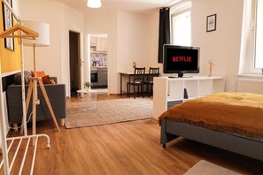 Una habitación de hotel moderna con una cama, un televisor mostrando 'NETFLIX', un escritorio con sillas, una kitchenette y un área de lavandería.
