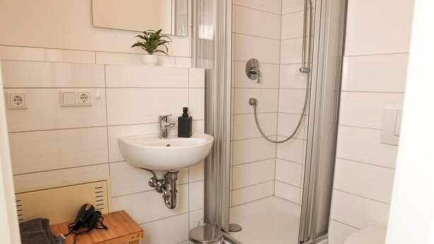 Een badkamer met een witte wasbak, een douche met glazen deur, een klein houten tafeltje en een prullenbak.